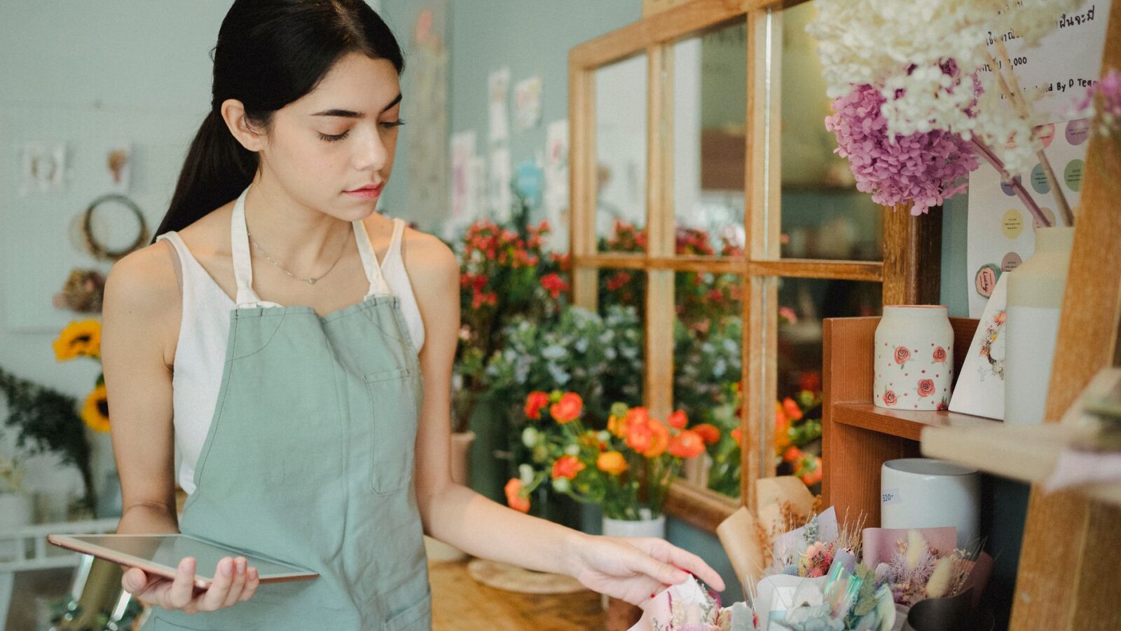Concentrated female florist with dark hair standing in floral shop with tablet near flowers in wrapper while checking order during work