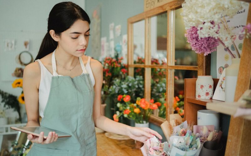 Concentrated female florist with dark hair standing in floral shop with tablet near flowers in wrapper while checking order during work
