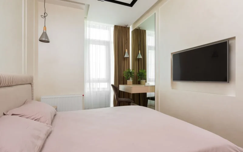 Large wall mirror opposite a bedroom window reflecting natural light into a room with pale walls and minimal furniture