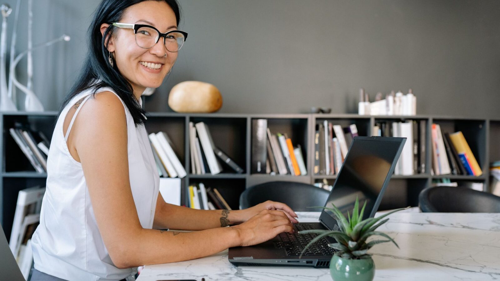 Cheerful businesswoman using laptop at a desk in a stylish office setup with bookshelves.