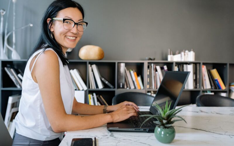 Cheerful businesswoman using laptop at a desk in a stylish office setup with bookshelves.
